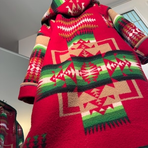 Op de afbeelding: Een rode wollen jas met een capuchon en een geometrisch patroon in groen, beige en wit. De jas heeft een zuidwestelijk ontwerp en hangt. De jas is middellang en heeft lange mouwen.