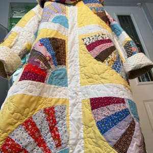 Puede incluir: Un abrigo acolchado hecho a mano con capucha. El abrigo presenta un diseño de patchwork con varias telas en amarillo, blanco, rojo, azul y estampados florales. El abrigo tiene un diseño largo y fluido con una textura acolchada.