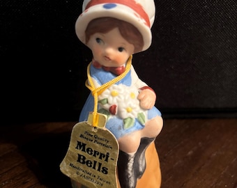 Puede incluir: Una figura de campana de porcelana de bizcocho de un niño con un gorro blanco con puntos rojos y azules, un vestido azul y zapatos negros. El niño sostiene flores blancas. Una cuerda amarilla sujeta una etiqueta que dice Fine Quality Bisque Porcelain Merri-Bells.