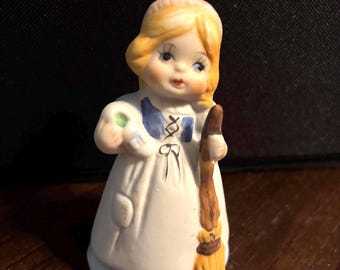 Puede incluir: Una figura de cerámica de una persona joven con cabello rubio que viste un vestido blanco con detalles en azul. La figura sostiene una escoba marrón en una mano y un pequeño objeto blanco con un centro verde en la otra.