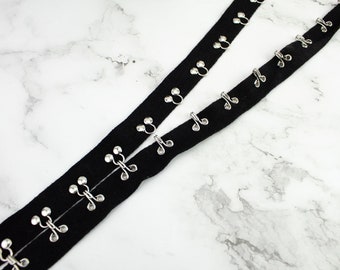 May include: Black elastic ribbon with silver metal hooks and loops. The ribbon is folded in half and the hooks and loops are visible on both sides.
