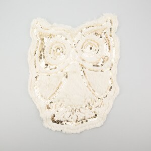 May include: A white owl applique with gold sequins. The owl is made of a soft, fuzzy fabric.