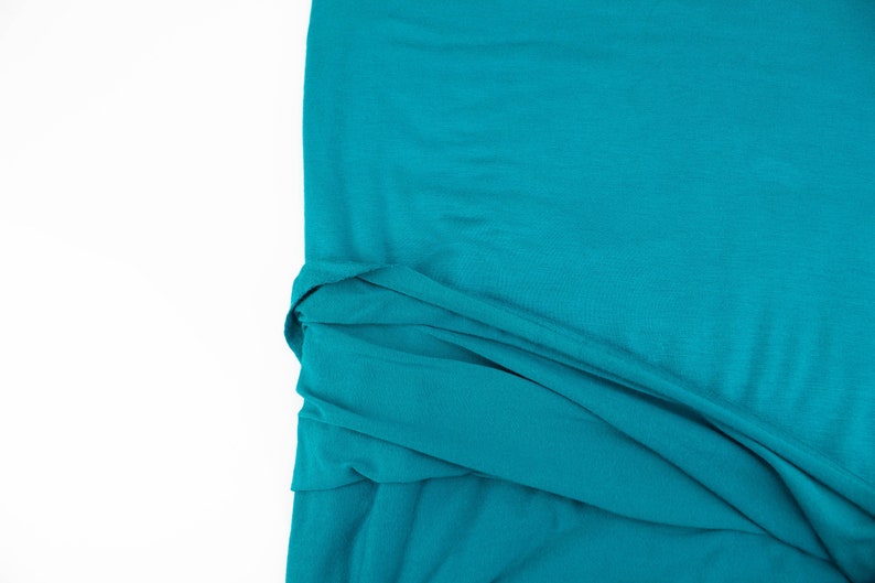 May include: A close-up of a teal fabric with a smooth texture. The fabric is folded and draped, creating soft folds and curves.