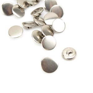 May include: A pile of silver metal buttons, some are round and some are slightly oval shaped. One button is laying on its side, showing the back of the button with a small hole in the center.