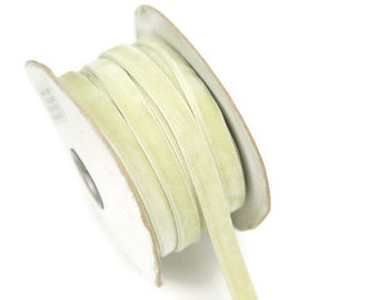 May include: A spool of pale green velvet ribbon. The ribbon is tightly wound on a cardboard spool, with a portion unwound and extending outwards. The ribbon has a soft, velvety texture and a subtle sheen, perfect for crafts and embellishments.