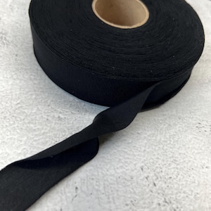 May include: A roll of solid black bias tape featuring a tightly wound spool with a cardboard center. The fabric ribbon has a smooth, matte texture and is shown with a single length of the tape extending outward from the main circular spool.