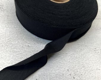 May include: A roll of solid black bias tape featuring a tightly wound spool with a cardboard center. The fabric ribbon has a smooth, matte texture and is shown with a single length of the tape extending outward from the main circular spool.