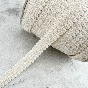 May include: A roll of off-white decorative trim with a scalloped edge. The trim is made of woven material with a textured, detailed pattern. The spool is partially visible, resting on a textured, light-colored surface.
