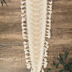 May include: Cream-colored table runner with a geometric design and tassel fringe. The runner features a central panel with a repeating pattern of triangles in a lighter shade, bordered by a cream-colored crochet trim and tassels.