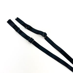 May include: Two black bra straps with adjustable metal sliders and rings. The straps are made of a smooth, woven material and are angled towards the top of the frame. The background is plain white.