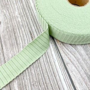 May include: A roll of light green elastic trim with a ribbed texture. The trim is rolled up and the end is unfurled, showing the texture.