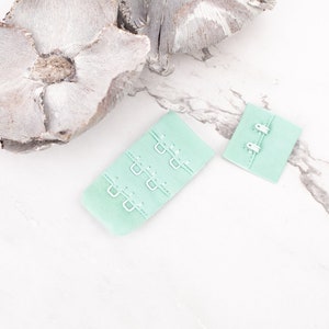 May include: Two light green bra hook and eye closures. One closure has three rows of hooks and eyes, the other has two rows.
