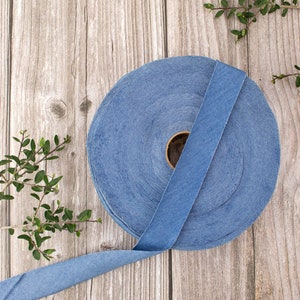 May include: A roll of blue denim ribbon on a wooden surface. The ribbon is rolled up and tied with a bow. There are green leaves in the background.