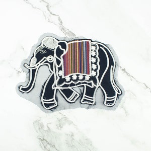 May include: A black elephant patch with white embroidered details and a colorful striped saddle blanket. The patch is on a gray mesh backing. The elephant design is a decorative element, suitable for clothing or accessories.