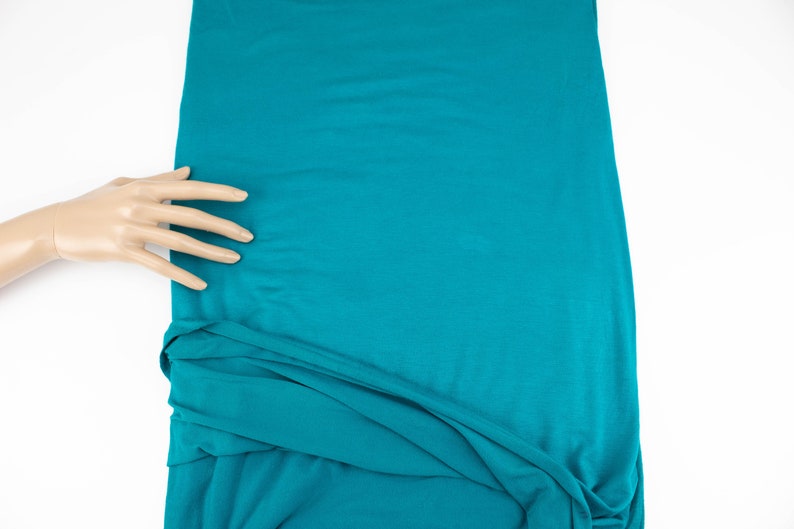May include: A teal blue fabric with a smooth texture. The fabric is folded and draped over a white surface.