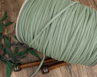 May include: A spool of sage green fabric ribbon. The ribbon is tightly wound on a white cardboard spool. The image is on a wooden surface with green leaves and two wooden spools in the background.