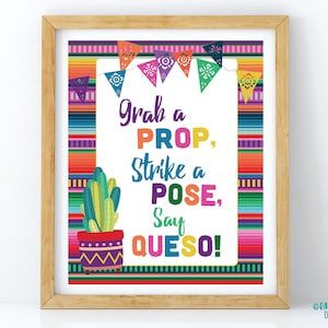 May include: A framed print with a colorful Mexican serape design. The print features the text "Grab a PROP, Strike a POSE, Say QUESO!" with a cactus illustration and decorative pennants. The frame is made of light brown wood.