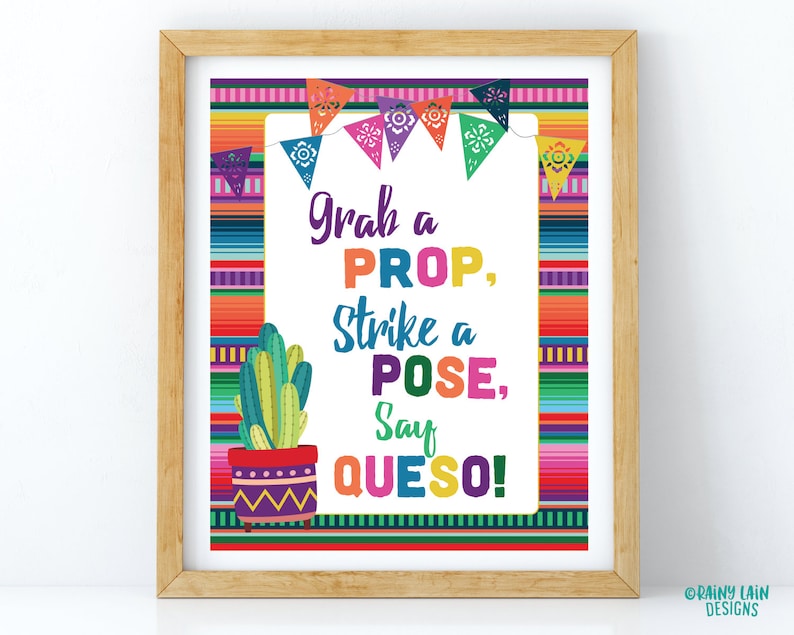 May include: A framed print with a colorful Mexican serape design. The print features the text "Grab a PROP, Strike a POSE, Say QUESO!" with a cactus illustration and decorative pennants. The frame is made of light brown wood.