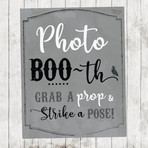 May include: A gray and white sign with the text "Photo Boo-th Grab a prop & Strike a pose!" in black lettering. A small black raven illustration is in the top right corner.