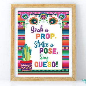 Può includere: Una stampa incorniciata con un design festoso. La stampa presenta il testo "Grab a PROP, Strike a POSE, Say QUESO!". Un cactus in vaso è a sinistra. La cornice è in legno.