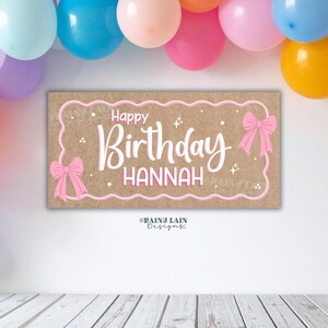 May include: A rectangular sign with the words "Happy Birthday Hannah" in white script, framed by a pink border and bows. The background is a light brown colour. Colourful balloons are visible in the background.