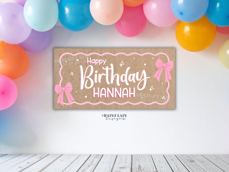 May include: A rectangular sign with the words "Happy Birthday Hannah" in white script, framed by a pink border and bows. The background is a light brown colour. Colourful balloons are visible in the background.