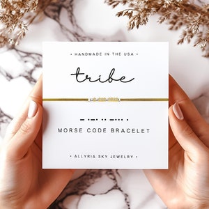 May include: A gold bracelet with a message in Morse code that reads "tribe". The bracelet is on a white card with the words "Handmade in the USA", "Morse Code Bracelet", and "Allryia Sky Jewelry".