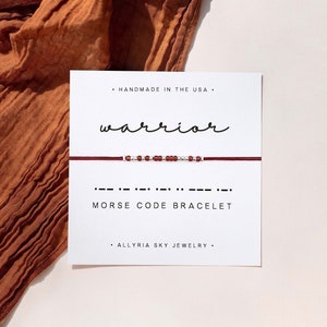 May include: A red string bracelet with silver beads spelling out "warrior" in Morse code. The bracelet is displayed on a white card with the words "Handmade in the USA", "Morse Code Bracelet", and "Allryia Sky Jewelry".