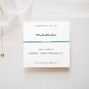May include: A white card with the text "HANDMADE IN THE USA" and "ALLYRIA SKY JEWELRY" on it. The card also has the word "mimi" written in cursive and the morse code for "mimi" written below it. The card is next to a green string bracelet with small beads.