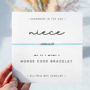 May include: A light blue bracelet with silver beads spelling out "niece" in Morse code. The bracelet is displayed on a white card with the text "Handmade in the USA", "niece", "Morse Code Bracelet", and "Allyria Sky Jewelry".