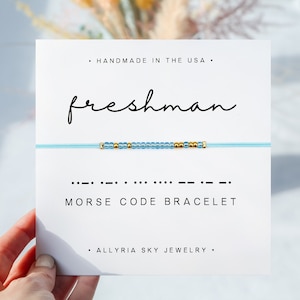 Peut inclure: Une carte blanche avec le texte "HANDMADE IN THE USA." et "freshman" en écriture cursive. Sous le texte se trouve un bracelet bleu perlé avec des perles dorées. Le bracelet est étiqueté "MORSE CODE BRACELET" et "ALLYRIA SKY JEWELRY."