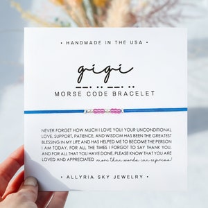 May include: A white card with the word "gigi" written in cursive and a line of dots and dashes representing the name in Morse code. The card also says "Morse Code Bracelet" and includes a message about love, support, and appreciation. The card is held in a hand, and a blue bracelet with pink beads is visible.