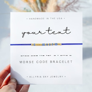 May include: A blue and gold beaded bracelet with the text "your text" and "MORSE CODE BRACELET" on a white card. The card also includes the text "HANDMADE IN THE USA" and "ALLYRIA SKY JEWELRY".