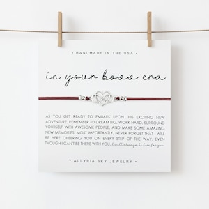May include: A red string bracelet with a silver charm that says "in your boss era". The bracelet is on a white card with the text "Handmade in the USA" and "Allryia Sky Jewelry".