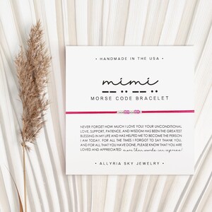 May include: A white card with the text "mimi" in black cursive script and ".. -- .." in black below it. The text "MORSE CODE BRACELET" is in black below the morse code. The card also includes the text "HANDMADE IN THE USA" and "ALLYRIA SKY JEWELRY". The card is sitting on a light brown surface.