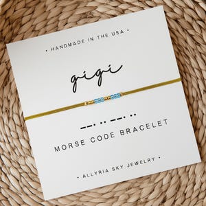 May include: A gold and blue beaded bracelet with the word "gigi" written in cursive on a white card. The card also says "Morse Code Bracelet" and "Handmade in the USA".