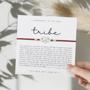 May include: A red string bracelet with a silver charm of two intertwined hearts. The bracelet is on a white card with the text "tribe" and a quote about the importance of finding your tribe.