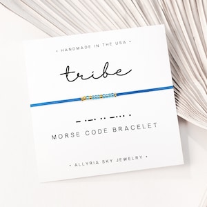 May include: A blue Morse code bracelet on a white card. The card reads "tribe" in cursive and "MORSE CODE BRACELET" in block letters. The bracelet has gold and blue beads. The card also says "HANDMADE IN THE USA."