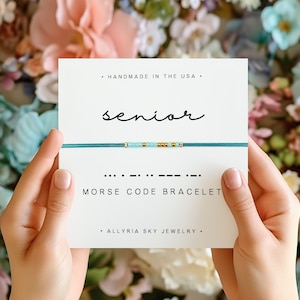 Peut inclure: Un bracelet en perles bleu turquoise et or avec le mot "senior" écrit sur une carte blanche. La carte indique également "Bracelet en code Morse" et "Fabriqué aux États-Unis".