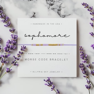 Peut inclure: Un bracelet en perles violettes et dorées avec le mot "sophomore" écrit en cursive au-dessus. Le bracelet est sur une carte blanche avec le texte "MORSE CODE BRACELET" et "HANDMADE IN THE USA" et "ALLYRIA SKY JEWELRY".