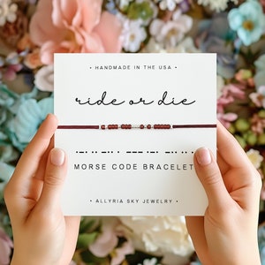 May include: A red and silver beaded bracelet with the phrase "ride or die" written in cursive on a white card. The card also includes the text "Morse Code Bracelet" and "Handmade in the USA".