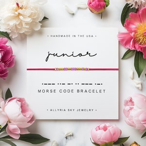 Peut inclure: Bracelet en cordon rouge avec des perles dorées et violettes qui épellent le mot "junior" en code Morse. Le bracelet est sur une carte blanche avec le texte "Fabriqué aux États-Unis", "Bracelet en code Morse" et "Allryia Sky Jewelry".