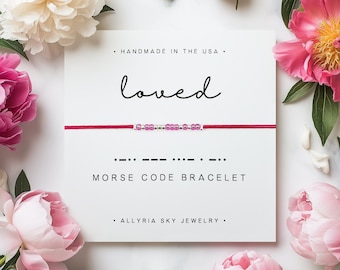 May include: A red string bracelet with silver and pink beads spelling out the word "loved" in Morse code. The bracelet is displayed on a white card with the text "loved" and "Morse Code Bracelet".