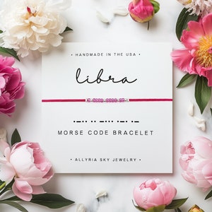 May include: A red string bracelet with pink beads spelling out "libra" in Morse code. The bracelet is on a white card with the text "HANDMADE IN THE USA", "libra", "MORSE CODE BRACELET", and "ALLYRIA SKY JEWELRY".