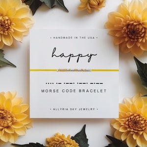 May include: A yellow and silver beaded bracelet with the word "happy" spelled out in Morse code. The bracelet is displayed on a white card with the words "Handmade in the USA", "Morse Code Bracelet", and "Allryia Sky Jewelry".
