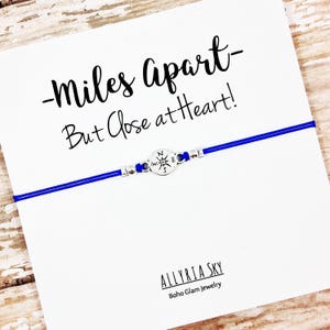 May include: A blue string bracelet with a compass charm and the text "Miles Apart - But Close at Heart!" and "ALLYRIA SKY Boho Glam Jewelry".