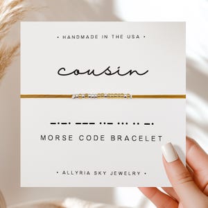 May include: A gold and silver beaded bracelet with a brown cord. The bracelet has the word "cousin" written on a card and the morse code for "cousin" written below it. The card also says "Handmade in the USA" and "Allryia Sky Jewelry".