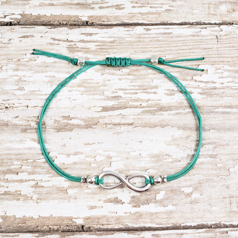 K&ouml;nnte beinhalten: Ein verstellbares Armband aus tealgr&uuml;nem Kordel mit einem silbernen Unendlichkeits-Anh&auml;nger und silbernen Perlen.