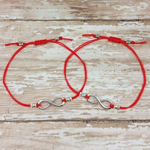 May include: Two red string bracelets with silver infinity symbol charms. The bracelets are tied together with a knot.