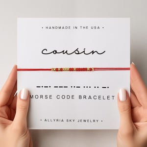 May include: Red string bracelet with gold beads spelling out "cousin" in Morse code. The bracelet is displayed on a white card with the text "Handmade in the USA", "Morse Code Bracelet", and "Allryia Sky Jewelry".